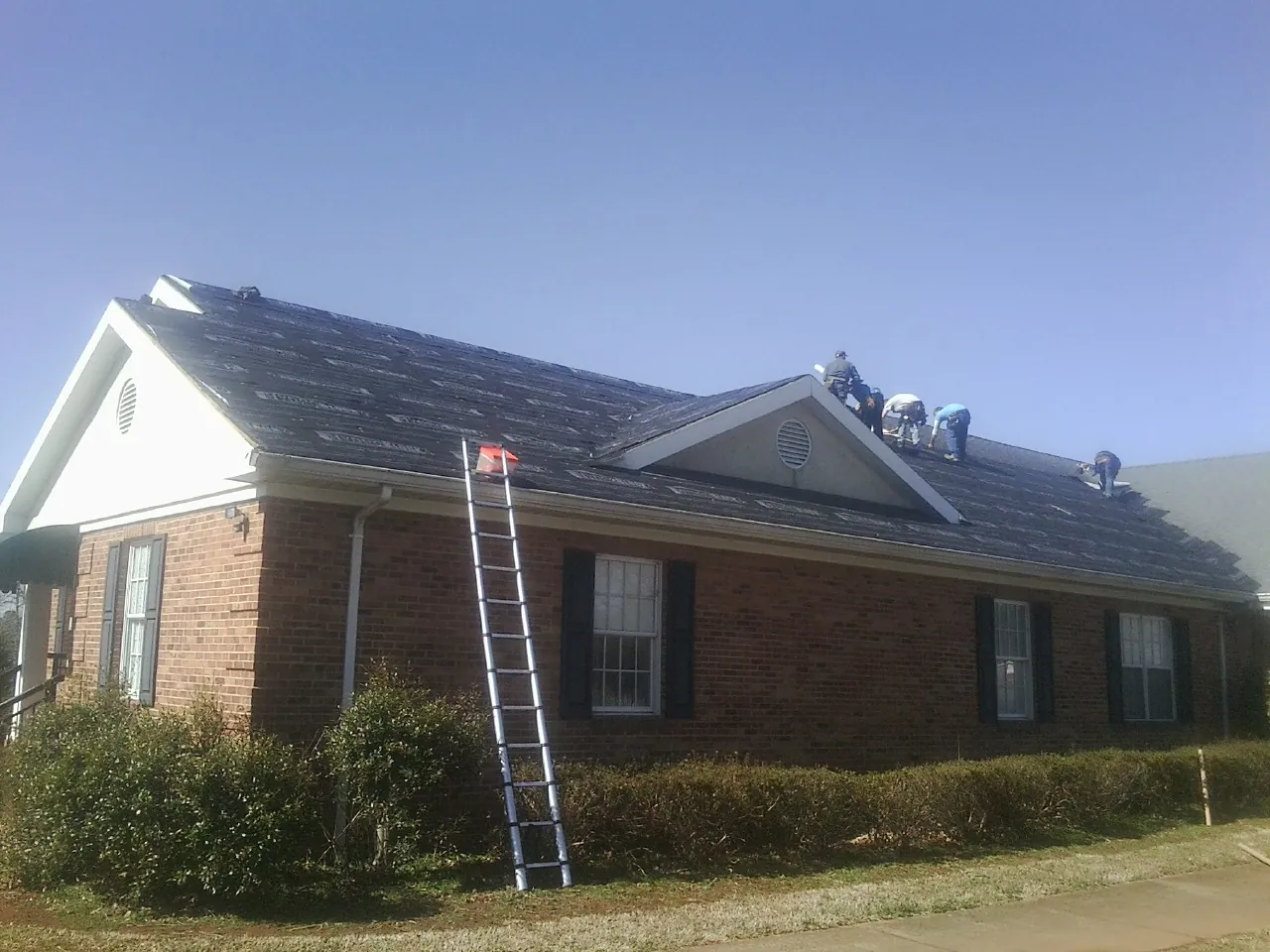 Crew tearing shingles off a brick ranch roof with a ladder set — Emergency Roof Repair work by our crew in Goodrich