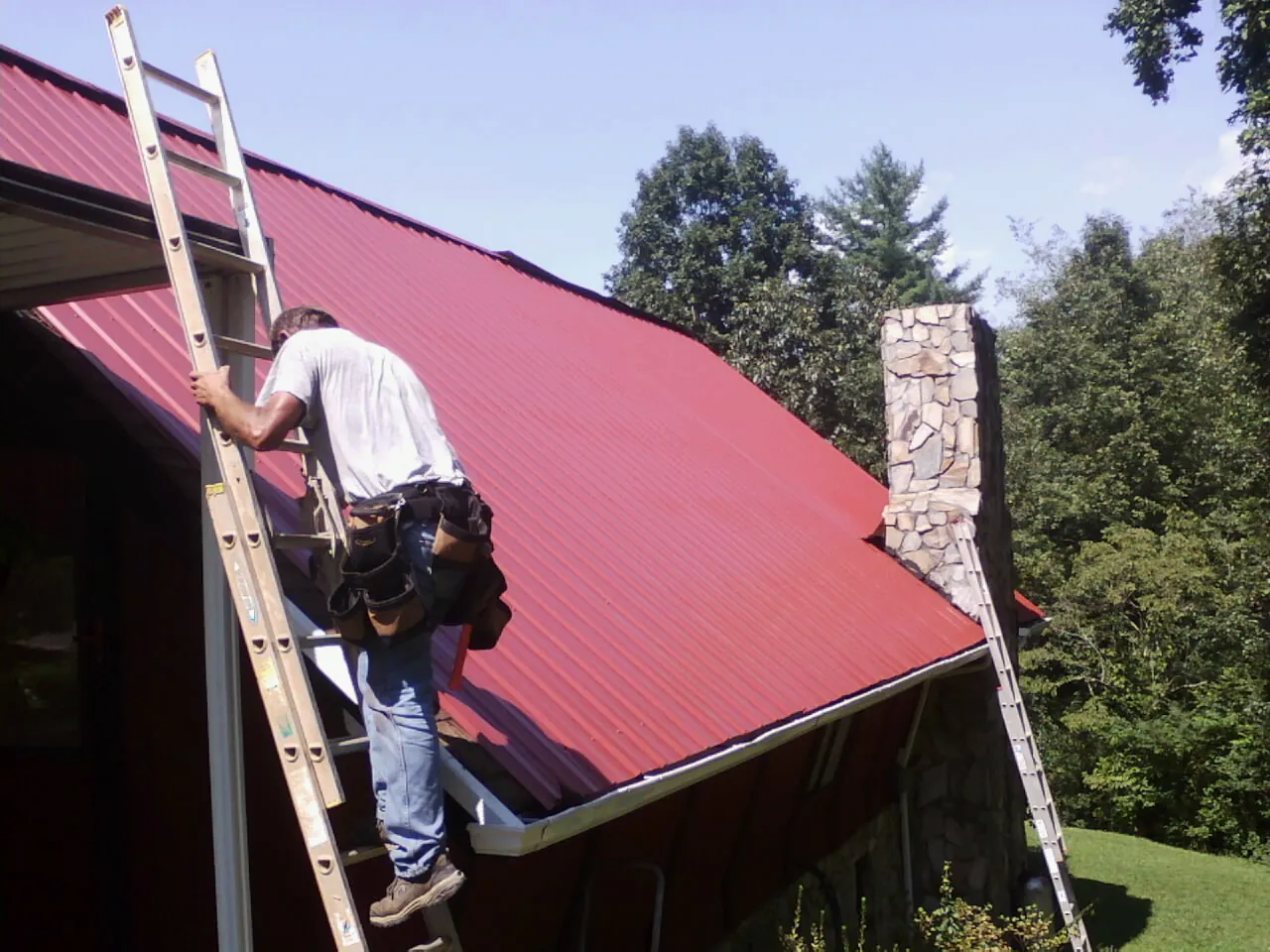 Roofer on a ladder beside a red metal roof and stone chimney — Soffit & Fascia Repair by Roofing Brothers in Goodrich